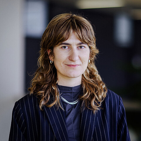 Porträt Harriet Meyer Eine Frau mit braunen Wellen, Ohrringen und gestreiftem Blazer blickt ernst in die Kamera.