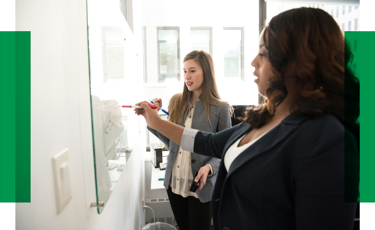 Two women are standing in front of a white board and work together.