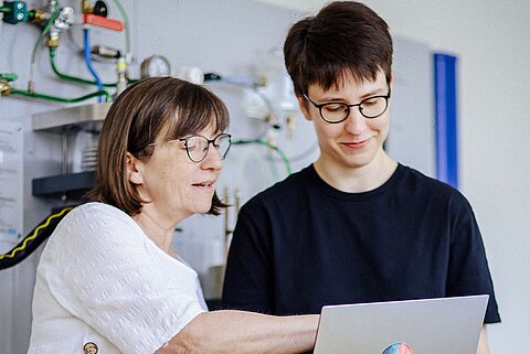 Technikbesprechung am Laptop Zwei Personen stehen vor einer technischen Wandinstallation. Eine zeigt auf den Laptop der anderen, beide tragen Brillen und wirken konzentriert.