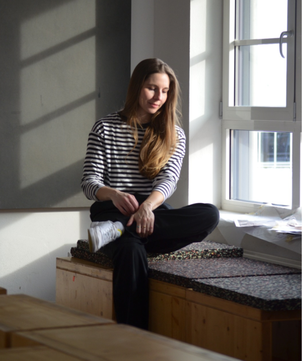 Andréa Hugen sitzt auf einer Holzkiste vor dem Fenster und schaut nach unten..