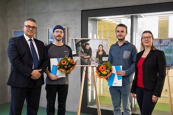 drei Männer und eine Frau stehen vor einem Bild auf einer Staffelei, zwei halten Urkunden und Blumen in den Händen