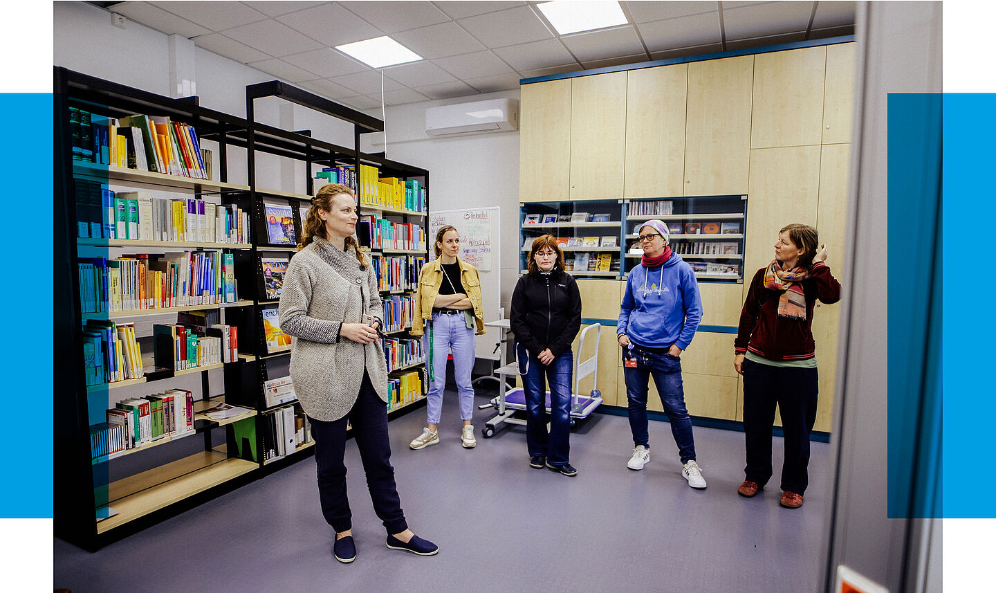 Fünf Personen stehen in einer modernen Bibliothek mit bunten Bücherregalen und hellen Holzschränken, im Gespräch miteinander.