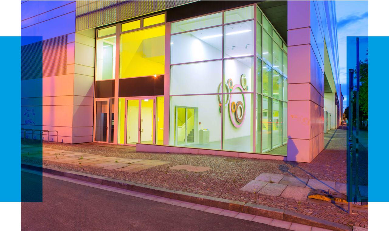 The photo shows a modern building facade at dusk. The building features a corner with large glass windows that allow a view into the brightly lit interior. The windows are tinted in a vibrant yellow, creating a striking contrast with the white and light gray cladding of the walls. Inside the building, minimalist white furniture and wall decorations in the form of abstract, circular shapes in shades of green are visible. The exterior lighting gives the building a warm glow. In front of the building, there is a walkway with paved stones and a small front garden area, suggesting an urban setting. On the right side of the image, there is a vertical blue bar, likely part of a graphic element, similar to the previous image.