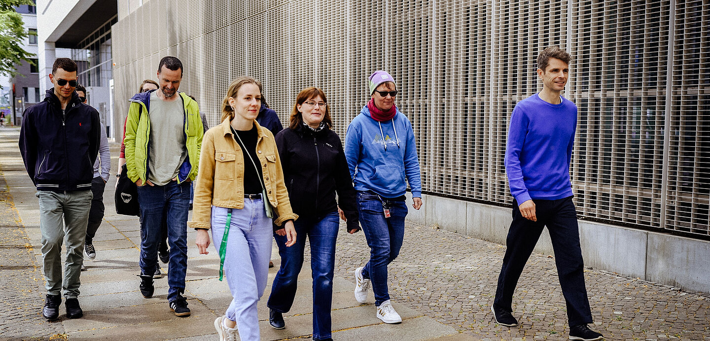 Gruppe Campustour 2024 Das Bild zeigt eine Gruppe von sechs Personen, die auf einem Gehweg entlang eines modernen Gebäudes spazieren. Die Gruppe geht in dieselbe Richtung und wirkt entspannt. Sie sind unterschiedlich gekleidet, passend zu einem eher kühlen Wetter.  Von links nach rechts:      Person 1: Trägt eine dunkle Sonnenbrille, eine dunkelblaue Jacke mit Reißverschluss, eine olivfarbene Hose und dunkle Schuhe. Die Hände sind in den Hosentaschen, der Kopf leicht nach unten geneigt, während die Person läuft.     Person 2: Trägt eine gelbe Jacke mit grünen Akzenten, darunter ein helles T-Shirt. Sie trägt eine graue Hose und dunkle Turnschuhe. Die Hände sind ebenfalls in den Taschen, der Kopf leicht nach unten geneigt.     Person 3: Trägt eine beige Jacke, ein schwarzes T-Shirt und eine hellblaue Jeans. Sie hat einen grünen Schlüsselanhänger, der an ihrer Jeans befestigt ist, und trägt weiße Sneaker. Ihre langen, hellbraunen Haare sind offen, und sie lächelt leicht, während sie mit einem energischen Gang läuft.     Person 4: Trägt eine schwarze Jacke und eine dunkle Jeans. Sie hat kurzes, dunkelbraunes Haar und trägt eine Brille. Die Hände sind nicht sichtbar, da sie hinter der Person 3 läuft. Ihr Gesichtsausdruck ist neutral.     Person 5: Trägt eine hellblaue Kapuzenjacke mit einem kleinen Motiv vorne, eine dunkle Jeans und weiße Sneaker. Sie hat eine Brille und trägt eine lila Mütze. Ein rotes Halstuch ist um ihren Hals gebunden, und sie trägt einen Schlüsselanhänger an der Jeans. Ihre Hände sind ebenfalls in den Taschen.     Person 6: Trägt einen lila Pullover und eine dunkelblaue Hose. Sie läuft etwas abseits von den anderen Personen auf der rechten Seite des Bildes. Ihr Gang ist entspannt, sie lächelt leicht, und ihre Hände sind locker an der Seite. Sie hat kurze, dunkelbraune Haare.  Der Hintergrund zeigt eine moderne Gebäudefassade mit vertikal verlaufenden Metallgittern und Fenstern. Der Gehweg besteht aus hellen und dunkleren Betonplatten, die in einem geometrischen Muster angeordnet sind. Im Hintergrund ist teilweise Laub auf dem Boden zu sehen. Es scheint tagsüber zu sein, mit natürlichem Licht, das die Szene beleuchtet.