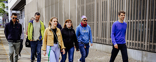 Gruppe Campustour 2024 Das Bild zeigt eine Gruppe von sechs Personen, die auf einem Gehweg entlang eines modernen Gebäudes spazieren. Die Gruppe geht in dieselbe Richtung und wirkt entspannt. Sie sind unterschiedlich gekleidet, passend zu einem eher kühlen Wetter. Von links nach rechts: Person 1: Trägt eine dunkle Sonnenbrille, eine dunkelblaue Jacke mit Reißverschluss, eine olivfarbene Hose und dunkle Schuhe. Die Hände sind in den Hosentaschen, der Kopf leicht nach unten geneigt, während die Person läuft. Person 2: Trägt eine gelbe Jacke mit grünen Akzenten, darunter ein helles T-Shirt. Sie trägt eine graue Hose und dunkle Turnschuhe. Die Hände sind ebenfalls in den Taschen, der Kopf leicht nach unten geneigt. Person 3: Trägt eine beige Jacke, ein schwarzes T-Shirt und eine hellblaue Jeans. Sie hat einen grünen Schlüsselanhänger, der an ihrer Jeans befestigt ist, und trägt weiße Sneaker. Ihre langen, hellbraunen Haare sind offen, und sie lächelt leicht, während sie mit einem energischen Gang läuft. Person 4: Trägt eine schwarze Jacke und eine dunkle Jeans. Sie hat kurzes, dunkelbraunes Haar und trägt eine Brille. Die Hände sind nicht sichtbar, da sie hinter der Person 3 läuft. Ihr Gesichtsausdruck ist neutral. Person 5: Trägt eine hellblaue Kapuzenjacke mit einem kleinen Motiv vorne, eine dunkle Jeans und weiße Sneaker. Sie hat eine Brille und trägt eine lila Mütze. Ein rotes Halstuch ist um ihren Hals gebunden, und sie trägt einen Schlüsselanhänger an der Jeans. Ihre Hände sind ebenfalls in den Taschen. Person 6: Trägt einen lila Pullover und eine dunkelblaue Hose. Sie läuft etwas abseits von den anderen Personen auf der rechten Seite des Bildes. Ihr Gang ist entspannt, sie lächelt leicht, und ihre Hände sind locker an der Seite. Sie hat kurze, dunkelbraune Haare. Der Hintergrund zeigt eine moderne Gebäudefassade mit vertikal verlaufenden Metallgittern und Fenstern. Der Gehweg besteht aus hellen und dunkleren Betonplatten, die in einem geometrischen Muster angeordnet sind. Im Hintergrund ist teilweise Laub auf dem Boden zu sehen. Es scheint tagsüber zu sein, mit natürlichem Licht, das die Szene beleuchtet.