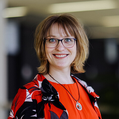 Porträt Dorothee Worringen Eine Frau mit blonden Haaren, Brille und roter gemusterter Kleidung steht lächelnd vor unscharfem Innenraum.