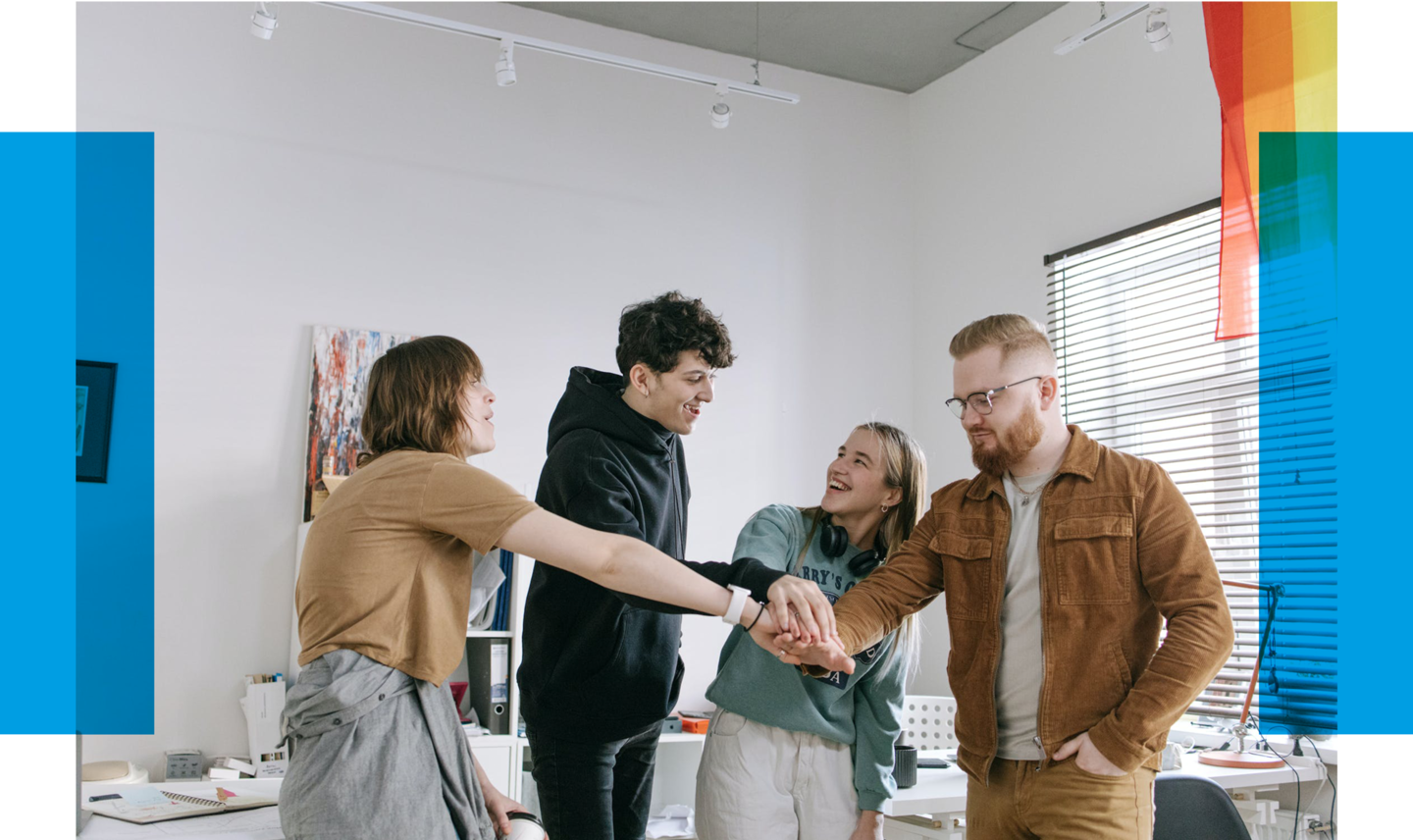 The photo shows four young adults in an office or seminar room, joyfully and amicably placing their hands together in the center of a circle. Two women and two men smile at each other, displaying signs of camaraderie or team spirit. The attire is casual; the men are wearing hoodies and shirts, while the women have casual tops, with one woman having headphones around her neck. The room is bright with artwork on the wall and a window letting in daylight. On the right side of the photo, part of a yellow and red flag is visible, suggesting an international context or a celebration. Yellow bars at the edges of the image might be part of a graphic web design.