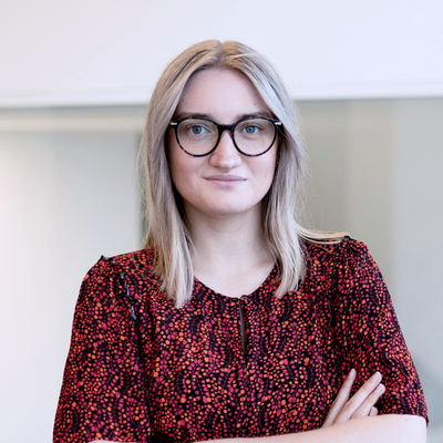 Frau mit blonden Haaren, schwarzer Brille und gemustertem Kleid steht mit verschränkten Armen vor heller Glaswand.