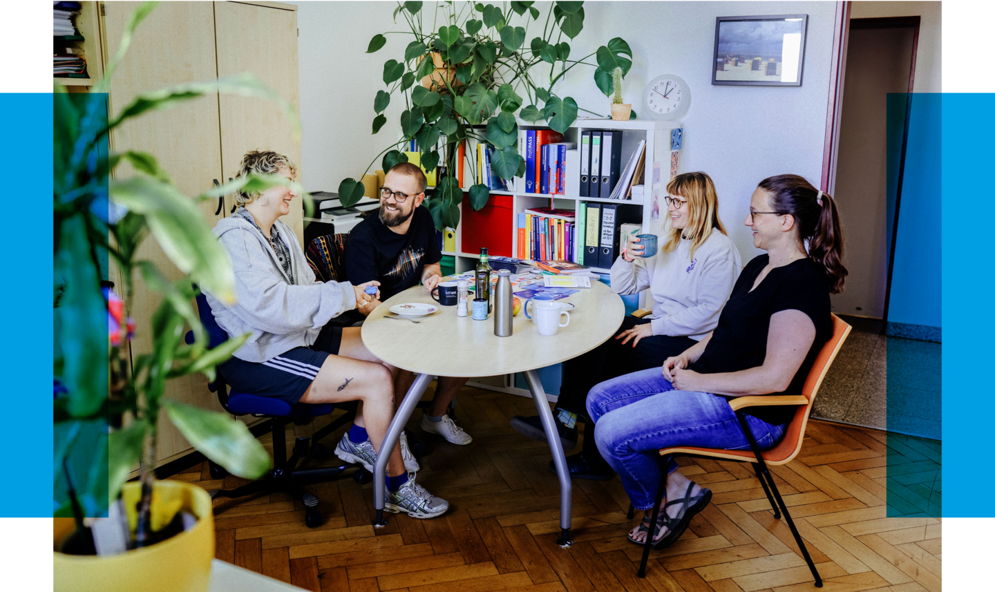 Headerbild Onboarding Vier Personen sitzen an einem runden Tisch in einem Büro, vor ihnen Getränke, im Hintergrund Pflanzen und ein Regal mit Ordnern.