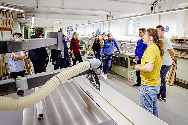 Das Bild zeigt eine Gruppe von Menschen in einer Werkstatt oder einem Labor. Eine Person in einem gelben T-Shirt steht rechts im Vordergrund und erklärt den Anwesenden etwas. Die Zuhörer, bestehend aus etwa acht Personen, sind in lockerer Kleidung und scheinen aufmerksam zuzuhören. Im Raum sind verschiedene Maschinen und Werkzeuge zu sehen, die auf eine handwerkliche oder technische Tätigkeit hinweisen. Die Atmosphäre wirkt geschäftig und konzentriert, und das natürliche Licht, das durch die Fenster einfällt, erhellt den Raum.