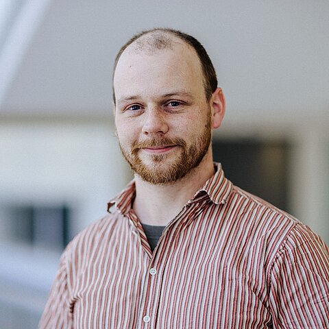 Porträt Vincent Weiss geschnitten Ein Mann mit lichtem Haar und Bart trägt ein rot kariertes Hemd und steht in einem hellen Innenraum, Blick zur Kamera.