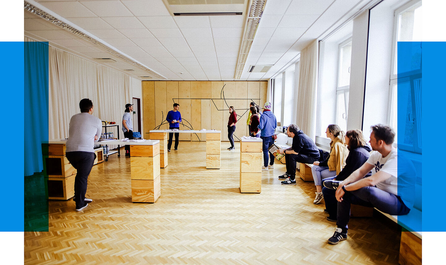 Das Bild zeigt eine Gruppe von Menschen in einem hellen Raum mit großen Fenstern und Holzparkettboden. Die Personen sind locker verteilt: Einige stehen an erhöhten Holztischen, während andere auf einer langen Sitzbank an der rechten Wand Platz genommen haben. Eine Person vorne links lehnt sich über einen Tisch, während eine andere Person in der Mitte des Raums steht und eine Präsentation zu leiten scheint. Die Wände sind teilweise mit Vorhängen und Holzelementen verkleidet, an denen geometrische schwarze Linien zu sehen sind, die eine dekorative Funktion haben. Der Raum wirkt wie ein kreativer Workshop- oder Meetingraum.