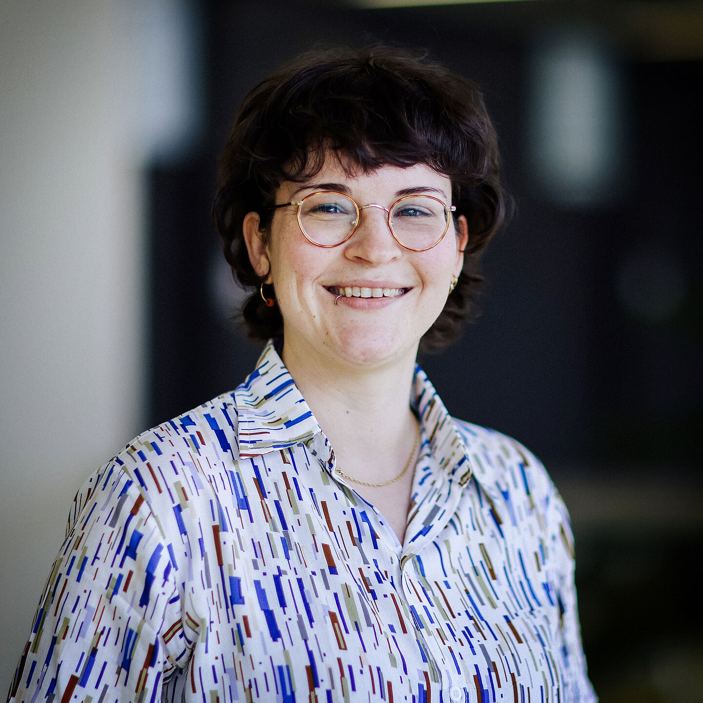 Nadine Glaser Frau mit dunklem, lockigem Haar, Brille und gemusterter Bluse steht lächelnd vor unscharfem Hintergrund.