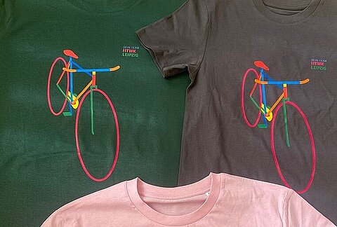 Stadtradeln 2024_Tshirts Drei T-Shirts in Pink, Grau und Grün mit identischem buntem Fahrradaufdruck liegen nebeneinander auf einer grünen Fläche.