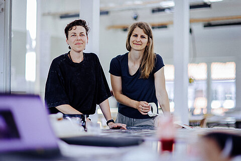 Zwei Frauen im Atelier Zwei Frauen stehen an einem Arbeitstisch in einem hellen Atelier. Eine hält Klebeband, beide schauen freundlich in die Kamera.