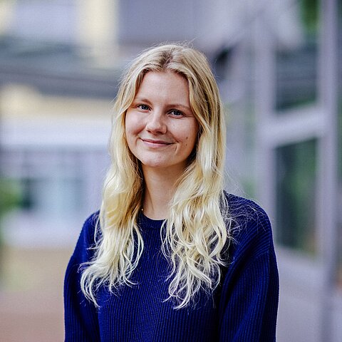 Porträt Tabea Rietz Eine junge Frau mit langen, gewellten blonden Haaren trägt einen dunkelblauen Pullover und steht lächelnd vor einem modernen Gebäude.