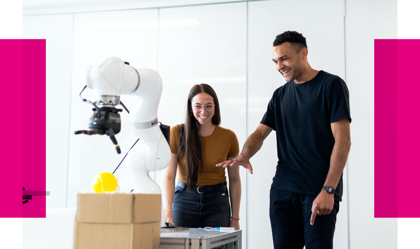 Auf dem Bild ist ein moderner, hell beleuchteter Raum zu sehen, in dem ein Mann und eine Frau lächelnd neben einem Tisch stehen, auf dem ein humanoider Roboter mit einem Greifarm platziert ist. Der Roboter scheint ein Objekt, das wie eine gelbe Kugel aussieht, zu manipulieren. Die Frau, die ein senfgelbes Oberteil und Jeans trägt, deutet mit ihrer offenen Hand auf den Roboter, während der Mann, in einem schwarzen T-Shirt und Jeans gekleidet, ihm mit einem interessierten Lächeln zuschaut. Im Hintergrund sind weiße Schränke und eine weiße Wand zu erkennen, was den Eindruck einer sauberen, technologieorientierten Umgebung vermittelt