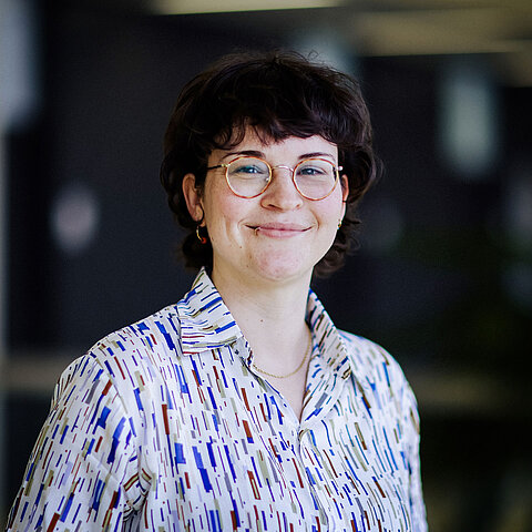 Porträt Nadine Glaser Eine junge Frau mit Brille, Kurzhaarschnitt und Lippenpiercing trägt ein bunt gemustertes Hemd, lächelt in die Kamera.