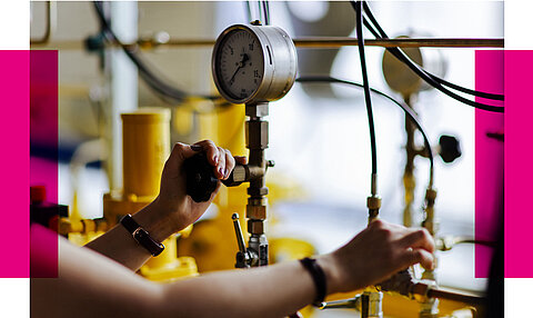Eine Person reguliert mit beiden Händen ein Ventil an einer technischen Anlage. Im Fokus steht eine Druckanzeige mit schwarzer Skala, die den aktuellen Druck misst. Die Anlage besteht aus gelben Rohren, schwarzen Schläuchen und Metallarmaturen. Die Umgebung ist leicht unscharf, wodurch die technischen Details betont werden.