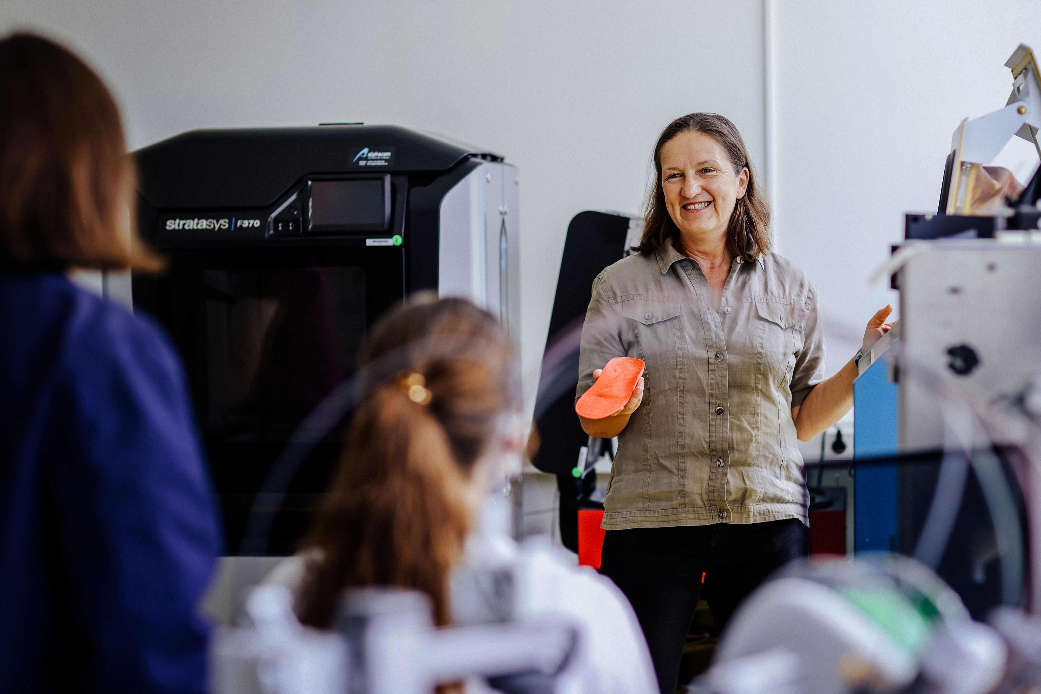Eine Frau steht vor einem 3D-Drucker, hält ein rotes Objekt in der Hand und spricht zu mehreren Personen im Raum.