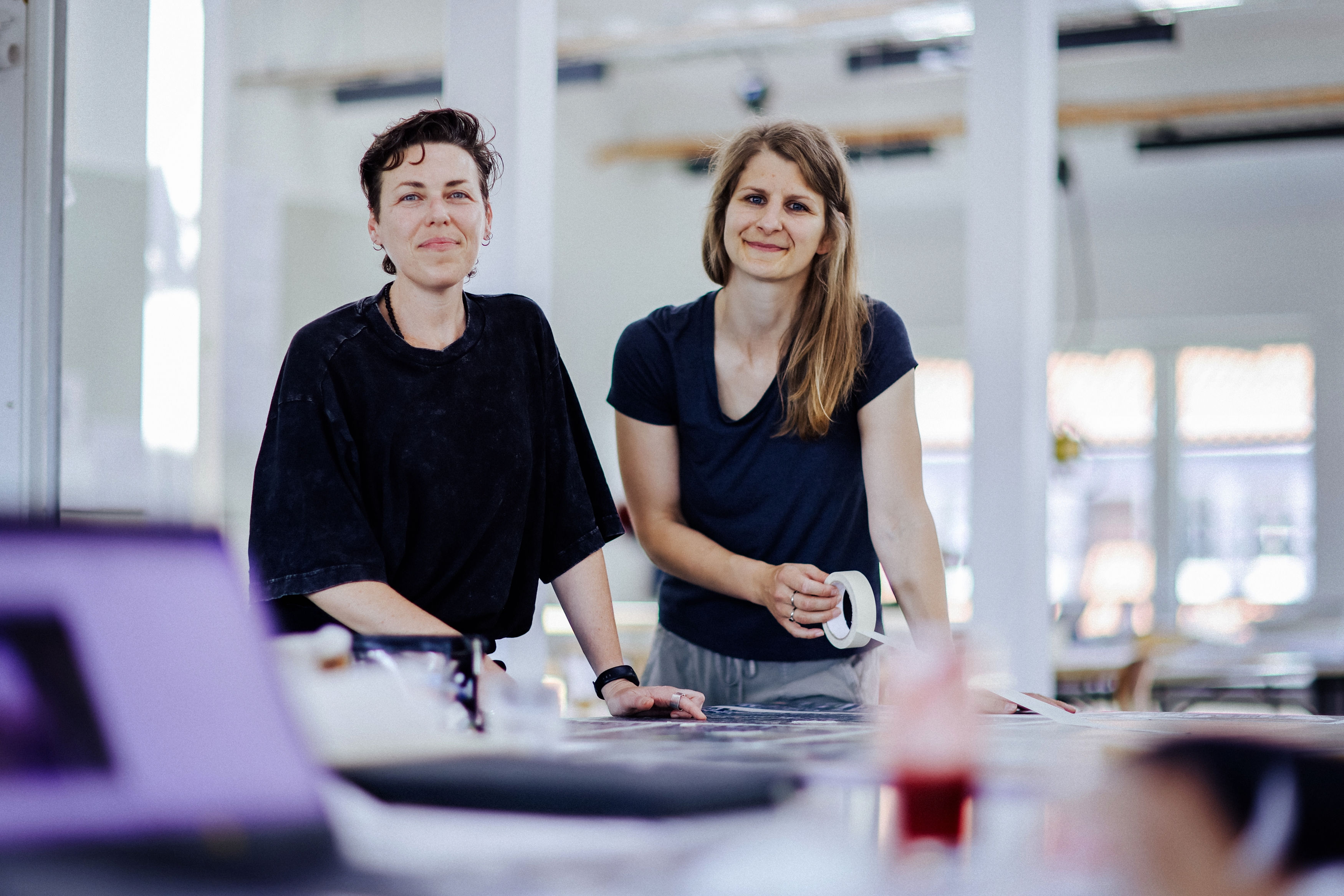 Zwei Frauen stehen an einem Arbeitstisch in einem hellen Atelier. Eine hält Klebeband, beide schauen freundlich in die Kamera.