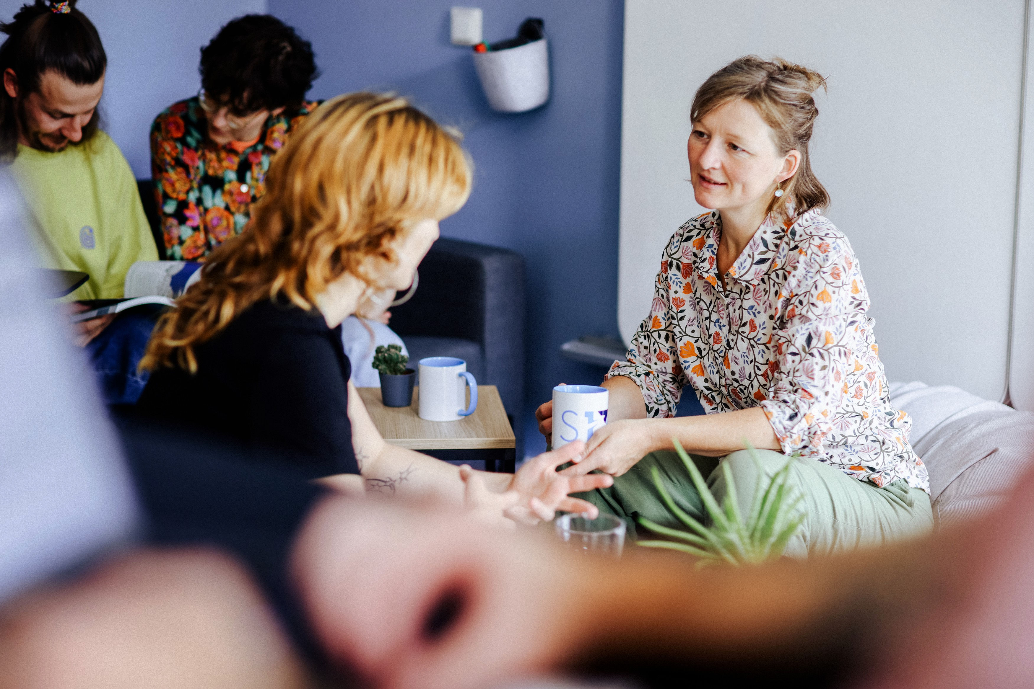 Mehrere Personen sitzen in einem Besprechungsrauum. Zwei Frauen im Vordergrund unterhalten sich, im Hintergrund lesen zwei Personen ein Buch.