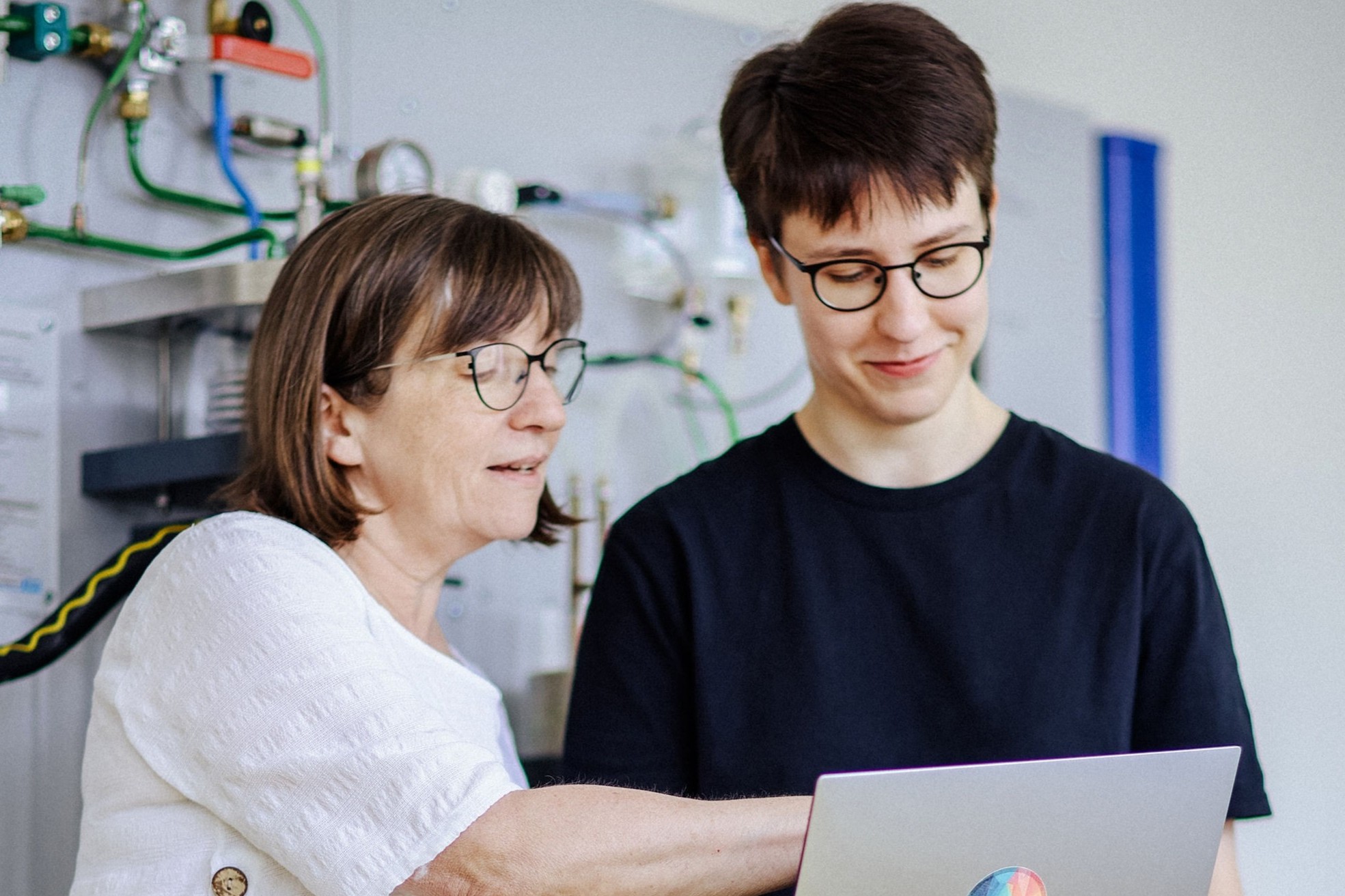 Zwei Personen stehen vor einer technischen Wandinstallation. Eine zeigt auf den Laptop der anderen, beide tragen Brillen und wirken konzentriert.