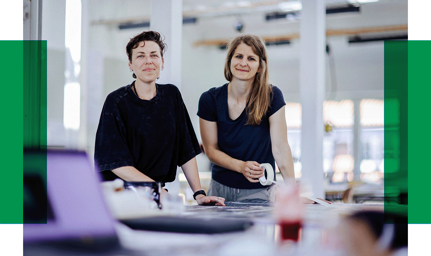 Zwei Frauen im Atelier Zwei Frauen stehen an einem Arbeitstisch in einem hellen Raum und lächeln in die Kamera.