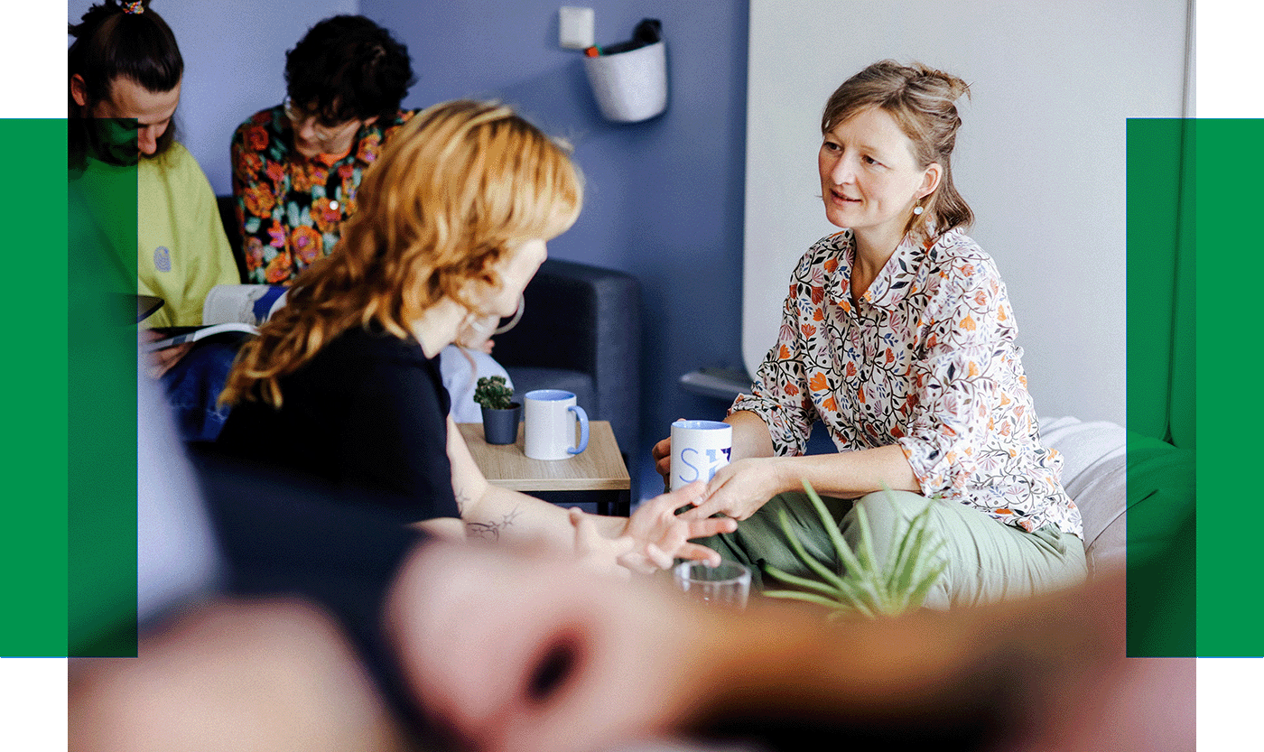 Gespräch in gemütlicher Runde Fünf Personen sitzen in einem hellen Raum, unterhalten sich und trinken Kaffee. Zwei Frauen sprechen sich direkt gegenüber.