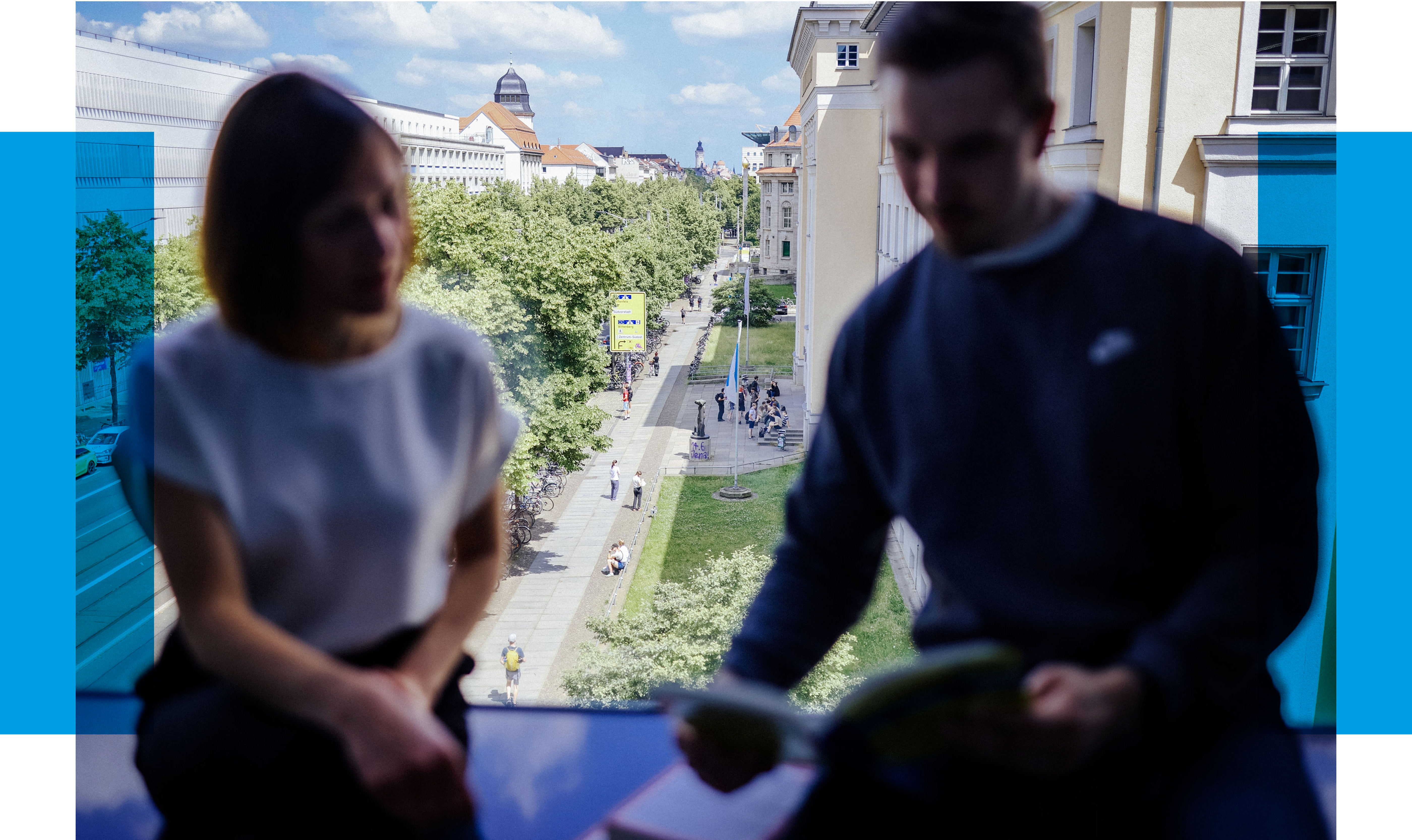  Eine Frau und ein Mann sitzen vor einem Fenster mit Blick auf eine belebte Straße und Bäume, beide lesen in einem Heft.