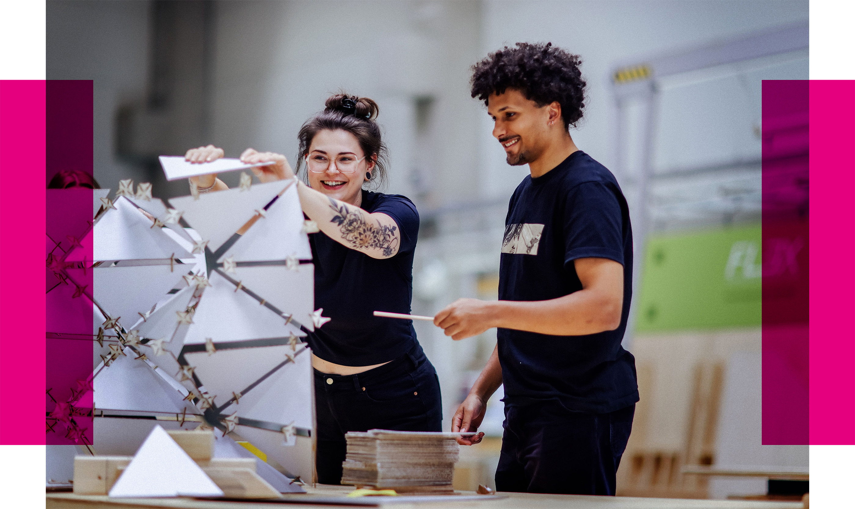 Zwei junge Personen arbeiten lachend an einem geometrischen Modell aus weißen, dreieckigen Elementen auf einem Werkstatttisch.