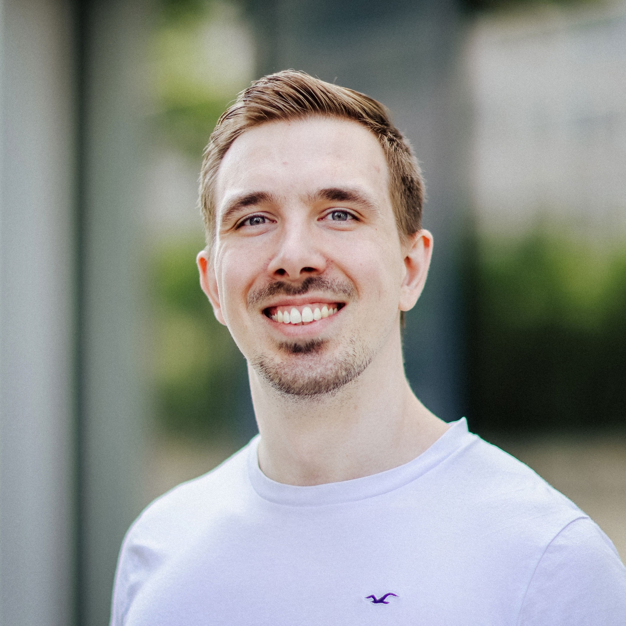 Porträt Jonas Gwdoz 2 Ein junger Mann mit kurzem Haar steht draußen, trägt ein weißes T-Shirt und lächelt in die Kamera, im Hintergrund unscharfe Architektur.