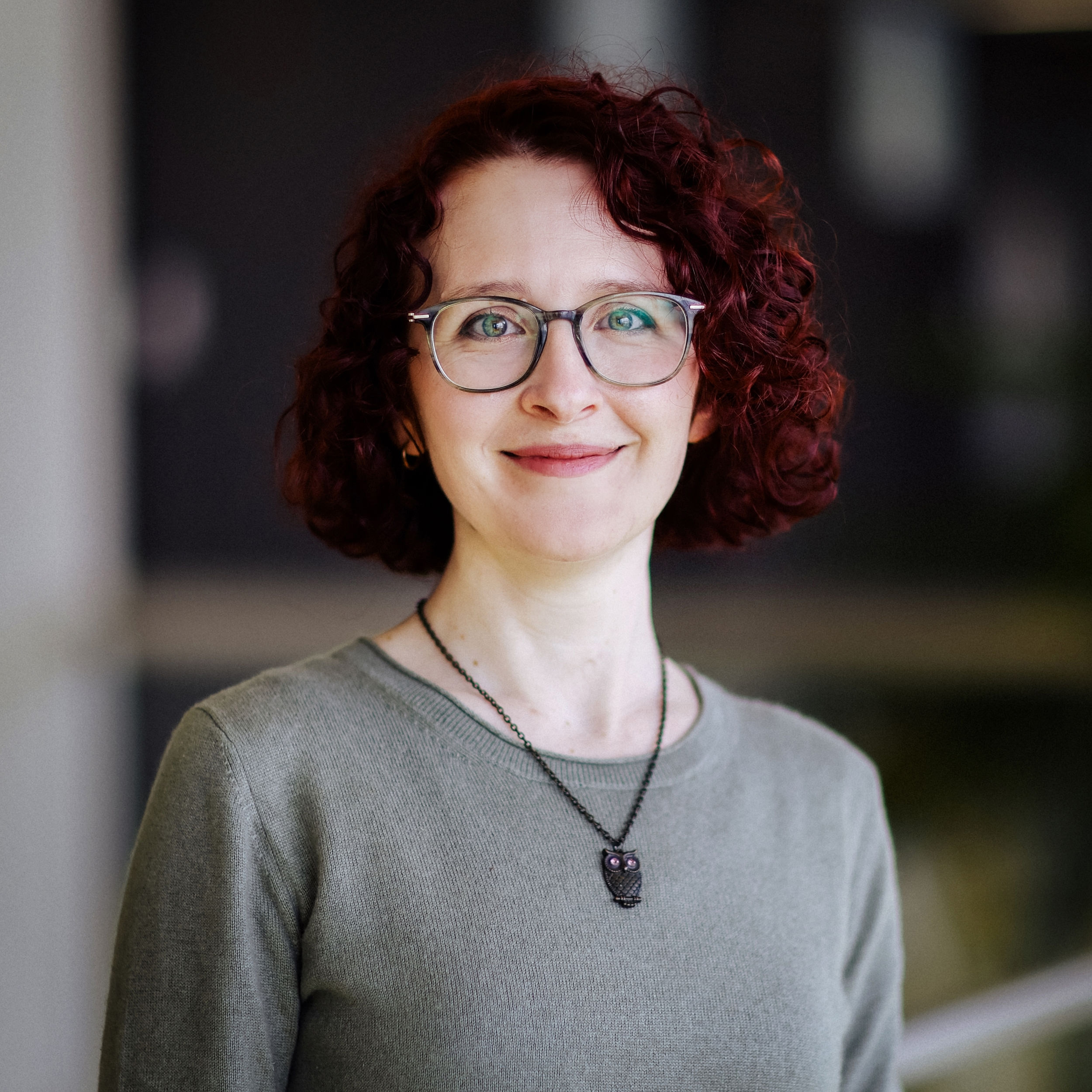 Porträt Therese Pagel Eine Frau mit roten Locken, Brille und grünem Pullover lächelt in die Kamera, neutraler Hintergrund.