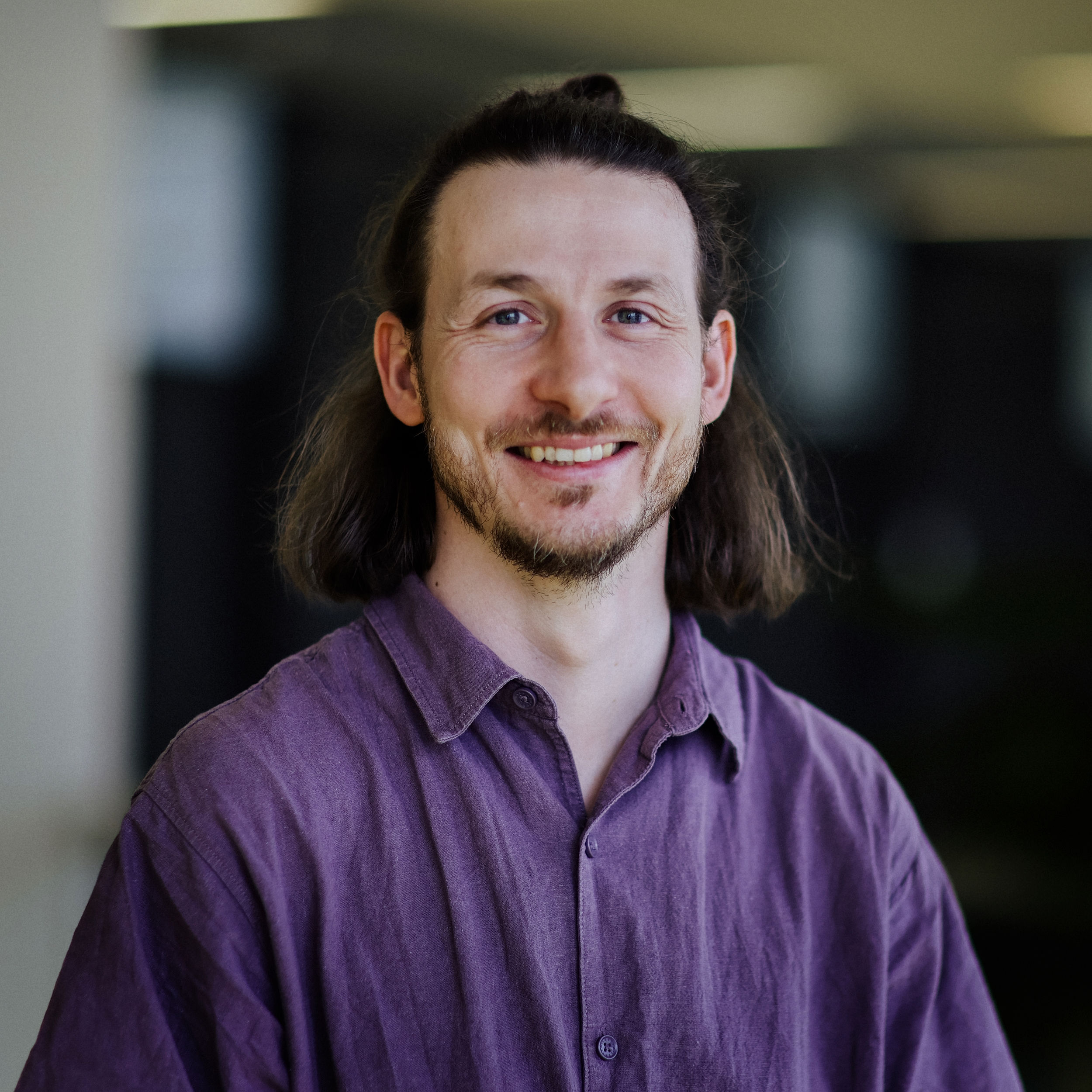 Porträt Sebastian Roscher Ein Mann mit schulterlangem Haar, Bart und lila Hemd steht lächelnd in einem Innenraum.
