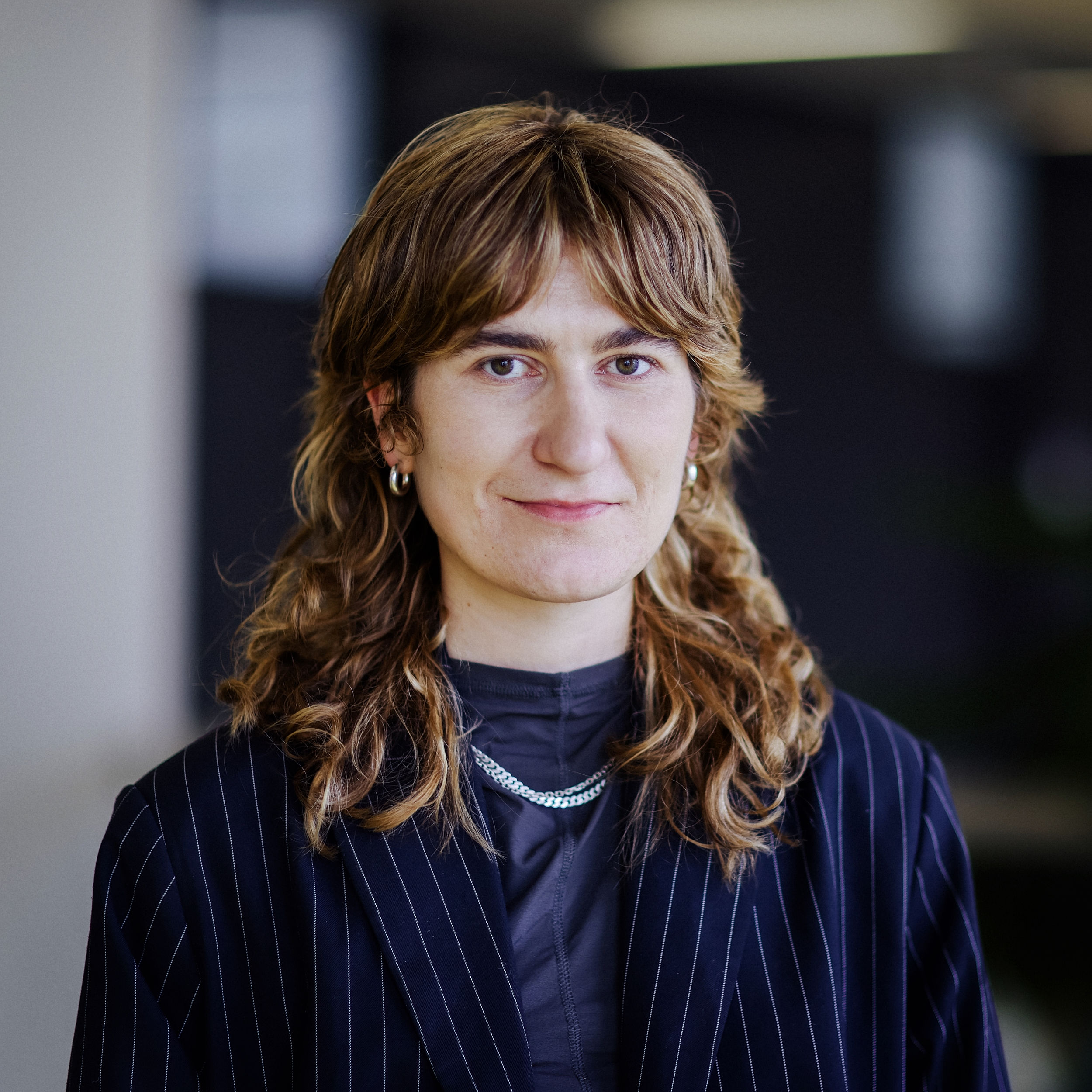 Porträt Harriet Meyer Eine Frau mit braunen Wellen, Ohrringen und gestreiftem Blazer blickt ernst in die Kamera.