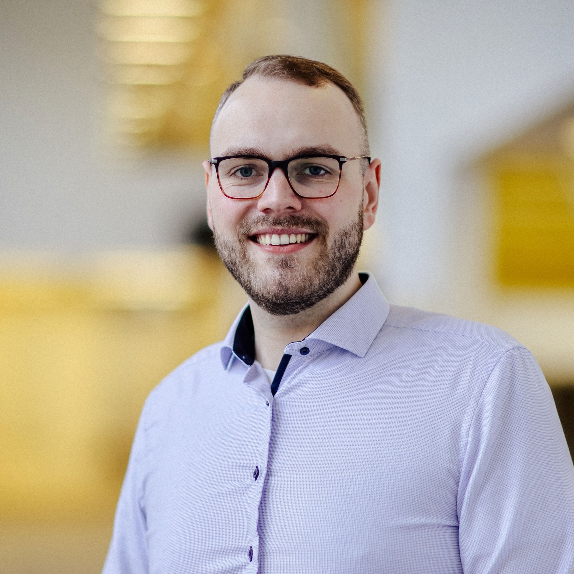 Porträt Lukas Hanisch geschnitten Ein Mann mit Brille, Bart und hellem gemusterten Hemd steht in einem modernen Innenraum und blickt lächelnd in die Kamera.